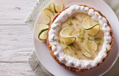 Fotografia em tons mais claros de uma torta inteira de limão com raspas e rodelas finas de limão. O chantilly está apenas na borda da torta, ao lado dela tem três fatias, que está sobre um prato grande e raso de vidro. Fotografia em tons mais claros de uma torta inteira de limão com raspas e rodelas finas de limão. O chantilly está apenas na borda da torta, ao lado dela tem três fatias, que está sobre um prato grande e raso de vidro.