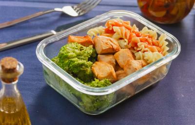 Fotografia mostra uma marmita em pote de vidro organizada com macarrão penne ao molho de tomate vibrante no canto superior direito, cubos de frango dourados ao meio, e brócolis verde intenso no espaço inferior.