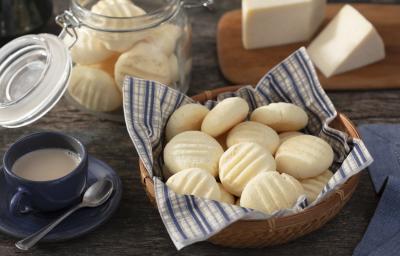 Fotografia em tons de azul em uma bancada de madeira escura, uma cesta de vime com um guardanapo de papel azul listrado com biscoitos de queijo dentro dele. Ao lado, uma xícara de café. Ao fundo, uma tábua de madeira com um pedaço de queijo. Fotografia em tons de azul em uma bancada de madeira escura, uma cesta de vime com um guardanapo de papel azul listrado com biscoitos de queijo dentro dele. Ao lado, uma xícara de café. Ao fundo, uma tábua de madeira com um pedaço de queijo.