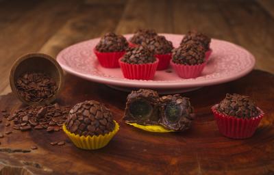 Fotografia em tons de amarelo e vermelho em uma bancada de madeira escura, um prato redondo rosa com brigadeiros em forminhas vermelhas em cima dele. Ao lado, brigadeiros em forminhas amarelas recheados com uva. Fotografia em tons de amarelo e vermelho em uma bancada de madeira escura, um prato redondo rosa com brigadeiros em forminhas vermelhas em cima dele. Ao lado, brigadeiros em forminhas amarelas recheados com uva.