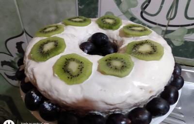 Foto da receita de bolo de cenoura confeitado servido com calda de chantilly, rodelas de kiwi cortadas por cima e uvas rosas para decorar em volta