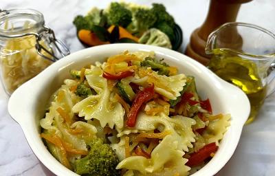 Foto em tons de amarelo da receita de farfalle com legumes servida em uma panela pequena de porcelana vermelha e branca. Ao fundo um vidro de azeite, um triturador de sal, uma porção de legumes e um pote de vidro com farfalle Foto em tons de amarelo da receita de farfalle com legumes servida em uma panela pequena de porcelana vermelha e branca. Ao fundo um vidro de azeite, um triturador de sal, uma porção de legumes e um pote de vidro com farfalle