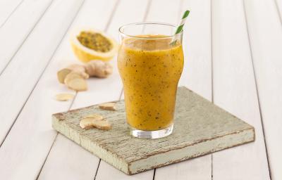 Fotografia em tons de amarelo em uma bancada de madeira branca, um paninho verde, uma tábua e madeira, um copo de vidro alto com o smoothie de manga com gengibre dentro dele. Ao fundo um pedaço de maracujá. Fotografia em tons de amarelo em uma bancada de madeira branca, um paninho verde, uma tábua e madeira, um copo de vidro alto com o smoothie de manga com gengibre dentro dele. Ao fundo um pedaço de maracujá.