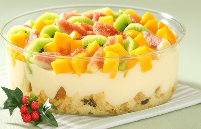 Fotografia em tons de amarelo, verde e vermelho em uma bancada de madeira verde clara, um pano branco com listras verdes, um recipiente de vidro com o pavê de panetone com frutas dentro dele.