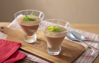 Fotografia de dois copinhos médios de vidro decorativos com a mousse sobre uma tábua de madeira retangular ao lado de duas colheres de sopa sobre um pano branco com listras vermelhas. Sobre as mousses tem folhas de hortelã. Fotografia de dois copinhos médios de vidro decorativos com a mousse sobre uma tábua de madeira retangular ao lado de duas colheres de sopa sobre um pano branco com listras vermelhas. Sobre as mousses tem folhas de hortelã.