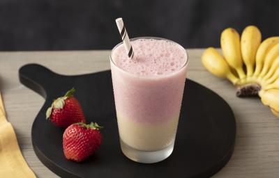 Fotografia em tons claros, em uma bancada de madeira bege escuro e ao lado, um pano amarelo. Ao centro, um prato redondo preto com um copo de smoothie morango e banana e um par de morangos ao lado. Ao fundo, um cacho de bananas. Fotografia em tons claros, em uma bancada de madeira bege escuro e ao lado, um pano amarelo. Ao centro, um prato redondo preto com um copo de smoothie morango e banana e um par de morangos ao lado. Ao fundo, um cacho de bananas.
