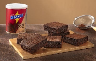 Foto da receita de brownie com nescau extra cacau servida em diversas porções sobre uma tábua de madeira, que acompanha uma peneira e uma embalagem de nescau ao fundo em cima de uma mesa de mármore escuro Foto da receita de brownie com nescau extra cacau servida em diversas porções sobre uma tábua de madeira, que acompanha uma peneira e uma embalagem de nescau ao fundo em cima de uma mesa de mármore escuro