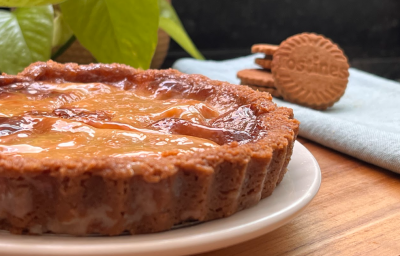 imagem mostra uma torta pequena sobre um prato branco. A torta em tons dourados com as bordas frisadas e um recheio caramelizado. Ao fundo um pano branco com 3 biscoitos Tostines e folhas verdes de uma planta.