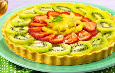 Fotografia em tons de verde e vermelho, com torta de frutas frescas fatiadas em prato verde sobre guardanapo verde com bolinhas brancas, potinho com mel ao fundo, tudo sobre bancada na cor cinza.