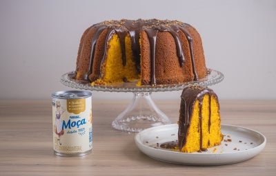 Foto da receita de Bolo de Cenoura com Cobertura de Brigadeiro, servido em um prato alto de vidro, com um pedaço cortado ao lado e, do lado esquerdo, uma lata de Leite Moça Foto da receita de Bolo de Cenoura com Cobertura de Brigadeiro, servido em um prato alto de vidro, com um pedaço cortado ao lado e, do lado esquerdo, uma lata de Leite Moça