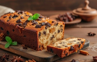 Fotografia de um bolo de frutas recém-assado, com crosta dourada e rica em passas e frutas secas. A fatia cortada revela a textura úmida, em um ambiente rústico com elementos decorativos como anis estrelado e canela.