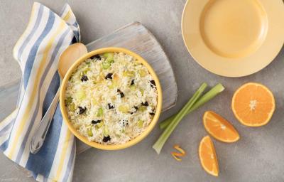 Fotografia em tons de azul e laranja em uma bancada de madeira cinza, um pano listrado amarelo e azul, uma tábua de madeira mesclada com um pote redondo grande amarelo com o arroz com laranja dentro dele. Ao lado, alho-poró e gomos de laranja. Fotografia em tons de azul e laranja em uma bancada de madeira cinza, um pano listrado amarelo e azul, uma tábua de madeira mesclada com um pote redondo grande amarelo com o arroz com laranja dentro dele. Ao lado, alho-poró e gomos de laranja.
