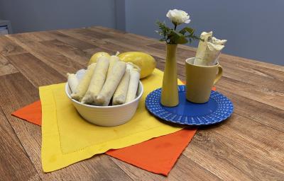 Foto da receita de Sacolé de Maracujá, servido em saquinhos, em tom amarelo, em uma travessa branca, sobre uma bancada de madeira decora com um tecido amarelo e outro laranja, ao lado há um prato azul redondo com um vaso e uma caneca amarelos