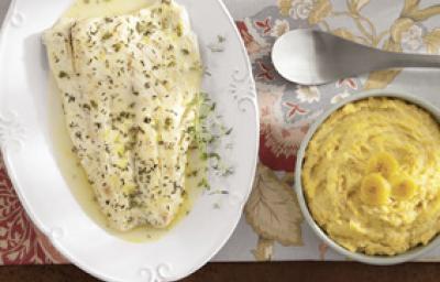 Fotografia em tons de amarelo em uma bancada de madeira com uma toalha florida, um prato oval branco com o peixe temperado com tomilho. Ao lado, um potinho branco com o purê de banana. Fotografia em tons de amarelo em uma bancada de madeira com uma toalha florida, um prato oval branco com o peixe temperado com tomilho. Ao lado, um potinho branco com o purê de banana.
