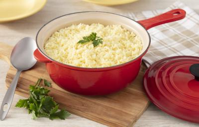 Fotografia em tons de vermelho de uma bancada branca, ao centro uma panela vermelha por fora e banca por dentro com arroz levemente amarelado. No topo, uma folha de salsinha decorando. Fotografia em tons de vermelho de uma bancada branca, ao centro uma panela vermelha por fora e banca por dentro com arroz levemente amarelado. No topo, uma folha de salsinha decorando.