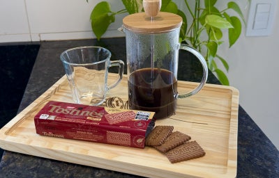 Foto da receita de Nescafé Gold com Especiarias, servido em uma prensa francesa, sobre uma bandeja de madeira e com um pacote de Biscoito Tostines Ginger na frente