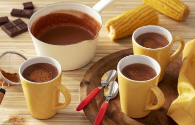 Fotografia em tons de amarelo em uma bancada de madeira de cor marrom. Ao centro, uma bandeja de madeira contendo 2 xícaras amarelas com 2 colheres ao lado. Ao fundo, há 2 espigas de milho, uma panela branca e uma xícara com chocolate quente. Fotografia em tons de amarelo em uma bancada de madeira de cor marrom. Ao centro, uma bandeja de madeira contendo 2 xícaras amarelas com 2 colheres ao lado. Ao fundo, há 2 espigas de milho, uma panela branca e uma xícara com chocolate quente.