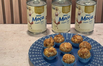 Docinhos cobertos com lascas de coco tostado em forminhas azuis, servidos em prato azul com Leite Moça ao fundo.