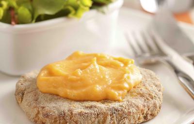 Fotografia em tons de vermelho em uma bancada de madeira com uma toalha listrada vermelha e bege, um prato com o hambúrguer de frango com cheddar derretido em cima. Ao fundo, um potinho com salada de alface. Fotografia em tons de vermelho em uma bancada de madeira com uma toalha listrada vermelha e bege, um prato com o hambúrguer de frango com cheddar derretido em cima. Ao fundo, um potinho com salada de alface.