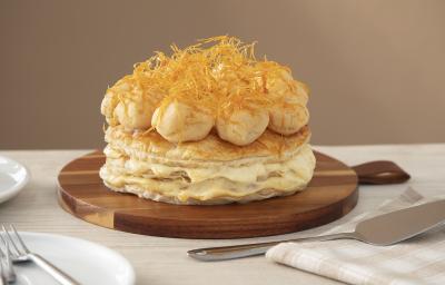 Foto da Receita de Gateau Saint Honoré. Observa-se a torta folhada sobre uma tábua redonda de madeira, num fundo e sobre uma superfície cor nude. Na ambientação, tem-se uma espátula de prata e um guardanapo quadriculado. Foto da Receita de Gateau Saint Honoré. Observa-se a torta folhada sobre uma tábua redonda de madeira, num fundo e sobre uma superfície cor nude. Na ambientação, tem-se uma espátula de prata e um guardanapo quadriculado.