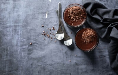 Fotografia vista de cima em tons escuros de dois copos de vidro com a sobremesa, finalizada com a ganache e raspas de chocolate. Ao redor tem duas colheres de sobremesa e um pano dobrado preto, sobre uma mesa de tom de cimento queimado escuro. Fotografia vista de cima em tons escuros de dois copos de vidro com a sobremesa, finalizada com a ganache e raspas de chocolate. Ao redor tem duas colheres de sobremesa e um pano dobrado preto, sobre uma mesa de tom de cimento queimado escuro.