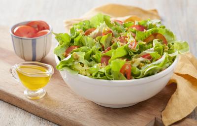 Fotografia em tons de  marrom e branco de uma bancada de madeira com uma tábua de madeira, sobre ela um recipiente branco redondo com salada. Ao lado um paninho amarelo, um recipiente com azeite e um recipiente com tomate.