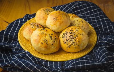 Foto aproximada de um prato amarelo com alguns pães de batata grande, decorados com gergelim. O prato está sobre um tecido azul quadriculado e numa bancada de madeira. Foto aproximada de um prato amarelo com alguns pães de batata grande, decorados com gergelim. O prato está sobre um tecido azul quadriculado e numa bancada de madeira.