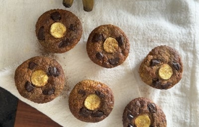 Fotografia em tons de branco com 6 muffins de banana ao centro. Os Muffins são finalizados com canela e uma fatia de banana por cima.