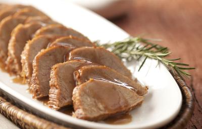 foto em tons marrom e branco. Uma mesa de madeira contém um cesto com um prato que comporta pedaços de carne fatiados.