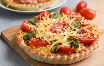 Imagem de uma quiche sobre uma tábua de madeira. A massa da quiche é dourada e crocante e ela está recheada com tomates-cereja, folhas de manjericão e lascas de queijo parmesão.