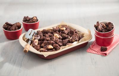 Fotografia em tons de vermelho em uma bancada de madeira clara com um refratário vermelho ao centro com castanhas crocantes com chocolate. Ao lado, potinhos vermelhos com pedaços de castanhas envoltas de chocolate.