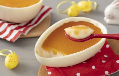 Fotografia em tons de vermelho, cinza e branco de uma bancada cinza com uma tábua marrom, sobre ela um paninho vermelho com um ovo de pascoa de pudim. Ao fundo mini ovinhos amarelos, uma tábua de madeira com um paninho listrado e um ovo de pudim. Fotografia em tons de vermelho, cinza e branco de uma bancada cinza com uma tábua marrom, sobre ela um paninho vermelho com um ovo de pascoa de pudim. Ao fundo mini ovinhos amarelos, uma tábua de madeira com um paninho listrado e um ovo de pudim.