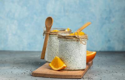 Fotografia em tons de azul com dois copos de vidro ao centro. Dentro de cada pote existe uma receita de Overnight Chia feita com laranja, chia e coco. Ao fundo existe uma laranja cortada ao meio