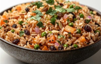 Em cima de uma mesa, tigela de porcelana redonda com porção de arroz com cebola, cenoura picada, uva-passa, ervilhas, decorado com coentro picado. Em cima de uma mesa, tigela de porcelana redonda com porção de arroz com cebola, cenoura picada, uva-passa, ervilhas, decorado com coentro picado.