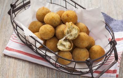 Fotografia em tons cinza, branco e dourado, mostrando bancada em tons de cinza com cestinha de arame contendo bolinhas de queijo fritas sobre papel toalha e guardanapo branco com listras vermelhas. Fotografia em tons cinza, branco e dourado, mostrando bancada em tons de cinza com cestinha de arame contendo bolinhas de queijo fritas sobre papel toalha e guardanapo branco com listras vermelhas.