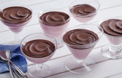 Fotografia em tons de chocolate e rosa, com seis taças contendo creme rosa e creme de chocolate, ao lado guardanapo azul e várias colheres, tudo sobre bancada em tom de rosa. Fotografia em tons de chocolate e rosa, com seis taças contendo creme rosa e creme de chocolate, ao lado guardanapo azul e várias colheres, tudo sobre bancada em tom de rosa.