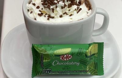Fotografia de uma xícara branca com um hot chocolate com chantilly e granulado por cima. Na frente da xícara tem um kitkat de menta, e está sobre um prato pequeno de vidro raso branco. Fotografia de uma xícara branca com um hot chocolate com chantilly e granulado por cima. Na frente da xícara tem um kitkat de menta, e está sobre um prato pequeno de vidro raso branco.