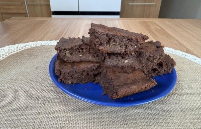 Fotografia de um prato raso azul sobre um paninho de tom bege apoiado em uma mesa de madeira. Sobre o prato tem alguns pedaços empilhados do brownie de abobrinha.