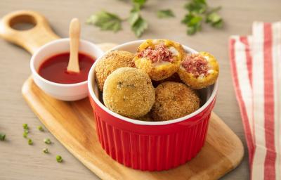 Fotografia em tons de amarelo, em uma bancada de madeira de cor marrom. AO centro, uma tábua de madeira com um recipiente vermelho contendo os bolinho de carne. Ao fundo, um pano listrado vermelho, um pote de katchup e folhas de salsinha espalhadas.