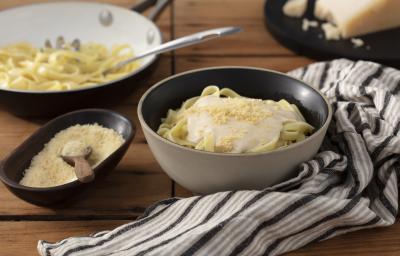 Fotografia em tons de marrom em uma bancada de madeira escura, um pano branco com listras marrom, um recipiente redondo fundo bege com o fettuccine com molho alfredo e queijo ralado por cima dentro dele. Ao lado, um potinho com queijo ralado.