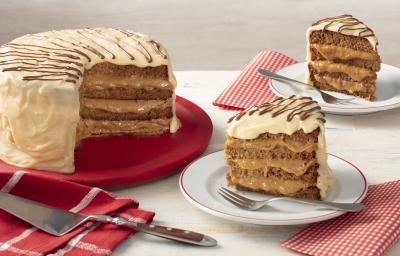 Fotografia em tons de vermelho em uma bancada cinza com paninhos vermelhos listrados e de bolinhas brancas. Ao centro, um prato branco com uma fatia do bolo de pão de mel com chocolate branco e à esquerda, um suporte grande vermelho com o bolo inteiro. Fotografia em tons de vermelho em uma bancada cinza com paninhos vermelhos listrados e de bolinhas brancas. Ao centro, um prato branco com uma fatia do bolo de pão de mel com chocolate branco e à esquerda, um suporte grande vermelho com o bolo inteiro.