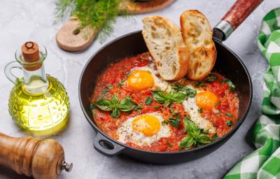 Fotografia em tons claros de uma receita de Shakshuka com pães por cima dentro de uma frigideira de ferro preta sobre uma mesa cinza. Ao lado da frigideira tem um recipiente com azeite, uma mini tábua de madeira e um moedor de pimenta de madeira. Fotografia em tons claros de uma receita de Shakshuka com pães por cima dentro de uma frigideira de ferro preta sobre uma mesa cinza. Ao lado da frigideira tem um recipiente com azeite, uma mini tábua de madeira e um moedor de pimenta de madeira.