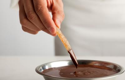 Fotografia de fruta seca afundando no chocolate amargo, que está em um recipiente de alumínio fundo. O qual está sobre uma mesa branca.