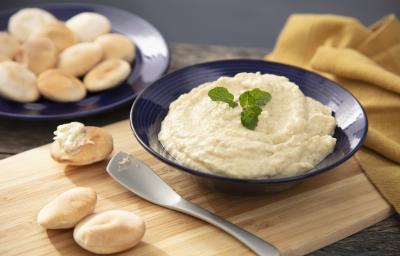 Fotografia em tons de amarelo em uma bancada de madeira, uma tábua de madeira, um prato azul redondo fundo com a babaganoush dentro e pães árabes ao lado. Ao fundo, mais pães e um pano amarelo. Fotografia em tons de amarelo em uma bancada de madeira, uma tábua de madeira, um prato azul redondo fundo com a babaganoush dentro e pães árabes ao lado. Ao fundo, mais pães e um pano amarelo.