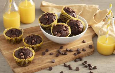 Fotografia em tons de amarelo em uma bancada de madeira com um pano bege, uma tábua de madeira ao centro, com os mini bolinhos de chocolate com gotas de chocolate em forminhas amarelas em cima da tábua e um pote branco com mais bolinhos.