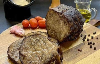 Foto em tons de marrom da receita de cupim na pressão servida em uma tábua de madeira com a peça de cupim cortada em algumas fatias e com tomates cerejas e pimentas. Ao fundo há uma jarra de vidro com azeite além de um recipiente preto com molho branco Foto em tons de marrom da receita de cupim na pressão servida em uma tábua de madeira com a peça de cupim cortada em algumas fatias e com tomates cerejas e pimentas. Ao fundo há uma jarra de vidro com azeite além de um recipiente preto com molho branco
