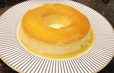 Foto aproximada de um pudim de leite condensado em tom amarelo, num prato redondo branco com decoração listrada. Na imagem há um selo em tom azul claro com o texto Receita do Consumidor. Foto aproximada de um pudim de leite condensado em tom amarelo, num prato redondo branco com decoração listrada. Na imagem há um selo em tom azul claro com o texto Receita do Consumidor.