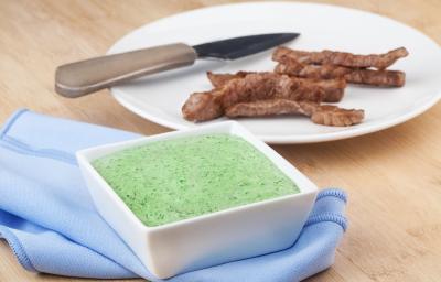 Fotografia em tons de azul e verde em uma bancada de madeira clara, um pano azul, um potinho quadrado branco com o patê cremoso de chimichurri dentro. Ao fundo, um prato branco redondo com tirinhas de carne para acompanhar o patê. Fotografia em tons de azul e verde em uma bancada de madeira clara, um pano azul, um potinho quadrado branco com o patê cremoso de chimichurri dentro. Ao fundo, um prato branco redondo com tirinhas de carne para acompanhar o patê.