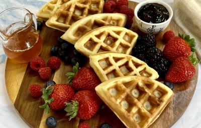 Foto em tons de bege e marrom da receita de waffle delícia servida em seis porções sobre uma tábua de madeira, com frutas vermelhas espalhadas pela tábua, além de um recipiente com geleia de frutas e vidro de mel