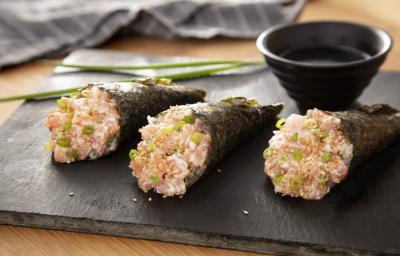Foto da receita de Temaki de Salmão. Observa-se 3 temakis em cima de uma tábua de preta e, atrás, um potinho com molho shoyu e um ramo de cebolinha.